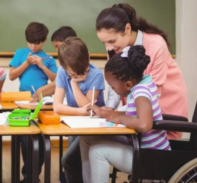 Imagem de duas crianças apoiada em uma mesa com uma mulher em pé vendo um caderno e duas crianças ao fundo desfocada.