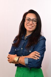 Foto da professora da Aline Morais. Possui cabelos escuros e lisos, olhos castanhos e usa óculos de armação preta, veste uma camisa jeans azul e está com o braço cruzado, no pulso esquerdo tem um relógio dourado e uma pulseira. O fundo da foto é branco.