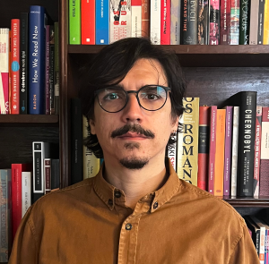 Antonio tem pele em tom claro, cabelos castanhos, curtos e lisos, cavanhaque também castanho e olhos castanhos. Usa óculos e camisa cáqui. Está à frente de uma estante com livros, com expressão séria.