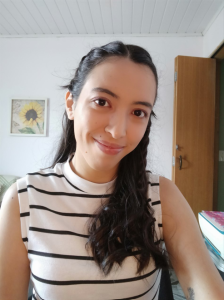 Elizabete tem pele em tom claro, cabelos castanhos, longos e ondulados e olhos castanhos. Usa camiseta branca com listras pretas horizontais. Está à frente de uma parede branca e uma porta, sorrindo.