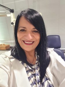 Foto da professora Eliana Cunha. Possui cabelo preto e liso soltos na altura do ombro, tem olhos castanhos e usa batom e uma camisa branca com detalhes azuis por baixo de um jaleco branco.