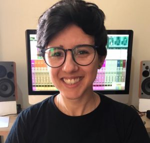 Laís tem pele em tom claro, cabelos castanhos, curtos e lisos e olhos castanhos. Usa óculos e camiseta preta. Está à frente de uma tela de computador, sorrindo.