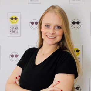 Cláudia tem pele em tom claro, cabelos loiros, compridos e lisos e olhos verdes. Usa uma camiseta preta. Está à frente de um backdrop com logo da Fundação Dorina, sorrindo de braços cruzados.