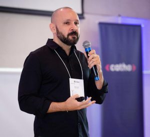 Foto do professor Rafael Públio que é careca e com barba, vestindo uma camisa preta. Ele segura um microfone na mão esquerda e gesticula com a direita, usa um crachá no pescoço e no fundo da foto há um banner da Catho.