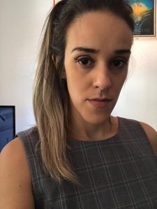 Tainá tem pele em tom claro, cabelos castanhos com luzes loiras, compridos e lisos e olhos castanhos. Usa uma blusa cinza axadrezada e está com os cabelos presos. Está à frente de uma parede branca, com expressão séria.