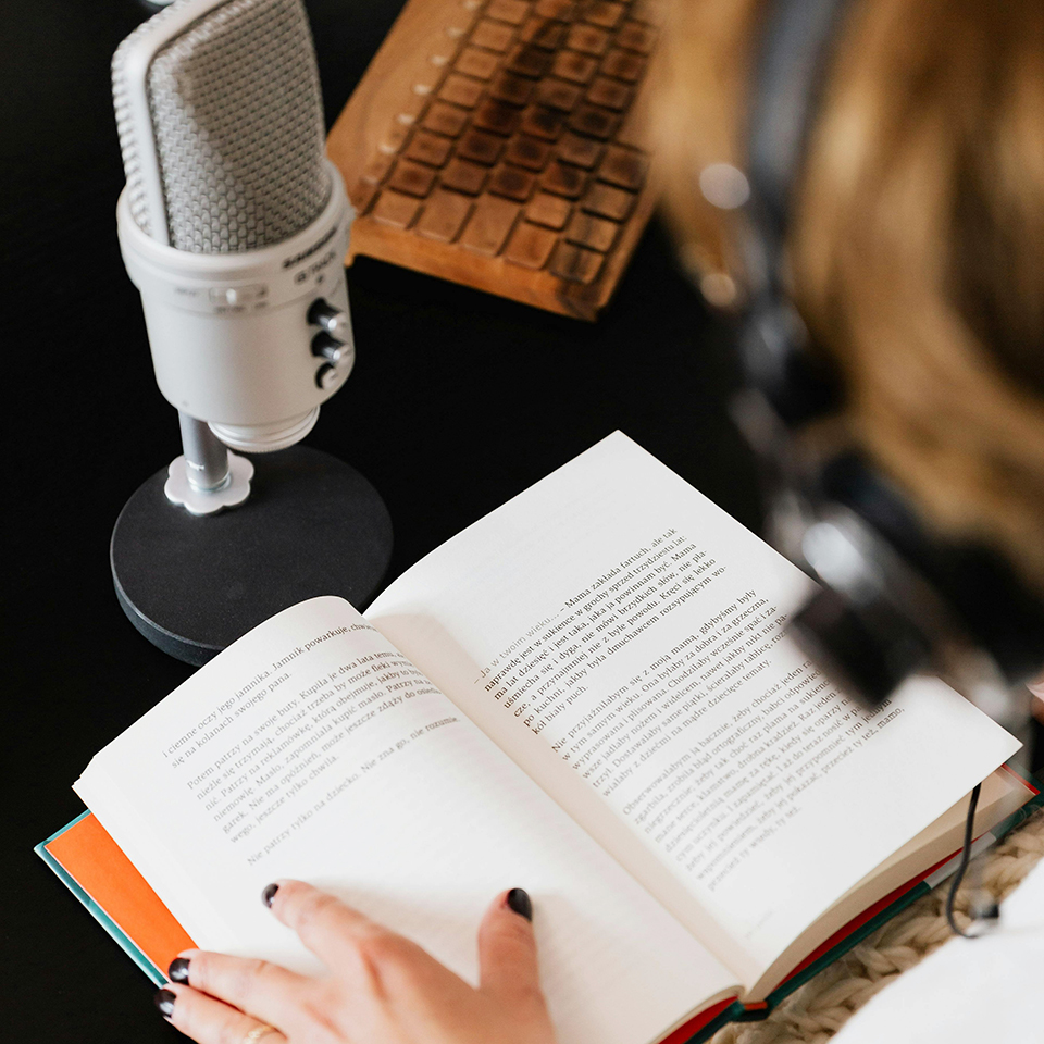 Imagem de um ponto de vista superior de um livro aberto, microfone de estúdio, teclado e uma pessoa com fone desfocada ao lado direito com a mão esquerda no livro.