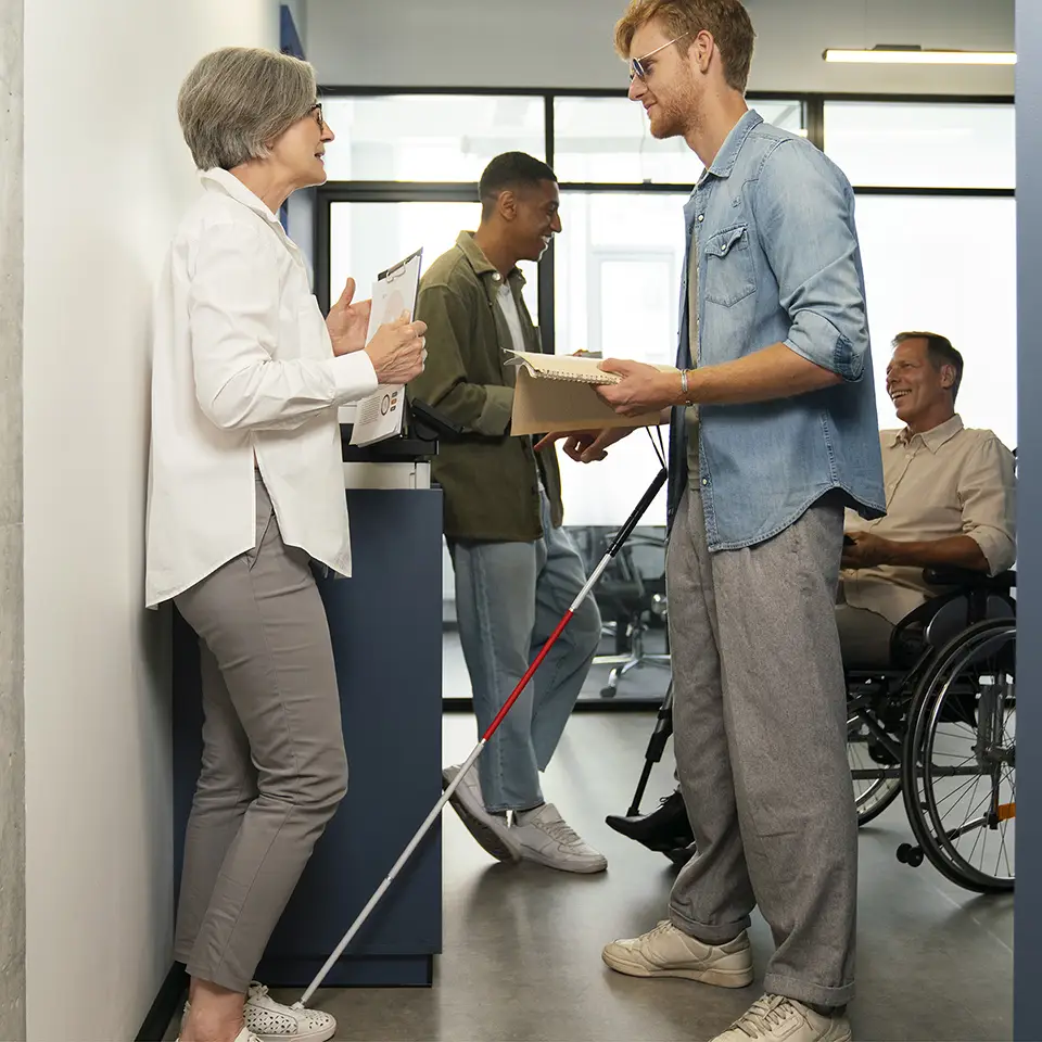 Foto de quatro pessoas de lado, duas a frente e duas ao fundo. À esquerda, uma mulher em pé com cabelos grisalhos e curtos, vestindo uma camisa branca e calça cinza. À direita um homem loiro de barba rala, veste uma camisa azul, uma calça cinza e usa uma bengala. No fundo há um homem em pé e um homem de cadeira de rodas.