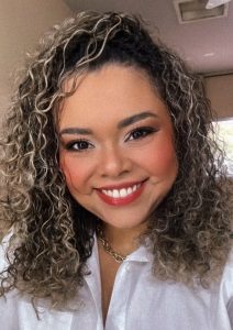 Foto da Professora Barbara que tem olhos castanhos, possui lábios e nariz finos, cabelos cacheados, com mechas loiras e usa maquiagem. Veste uma camisa branca e um colar no pescoço